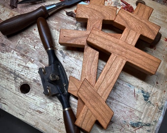 May include: Two handcrafted wooden Orthodox crosses with a smooth, natural finish, featuring a three-bar design. The crosses are positioned alongside various woodworking tools, including a metal spokeshave and several chisels with wooden handles.
