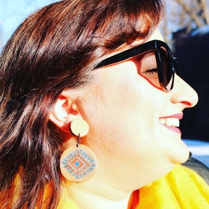 Peut inclure: Une femme portant des lunettes de soleil noires et une chemise jaune avec de grandes boucles d'oreilles rondes en bois avec un motif géométrique dans des tons de bleu, marron et blanc.