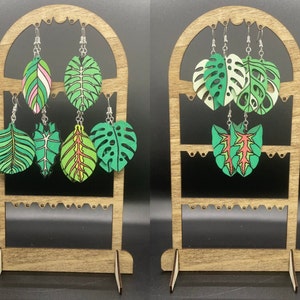 May include: A wooden display stand with two tiers holds a variety of leaf-shaped earrings. The earrings are made of wood and painted in shades of green, red, and white. The stand is a light brown color and has a decorative design.