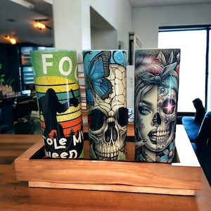 May include: Three stainless steel tumblers with different designs. The first tumbler has a green background with a black silhouette of a person holding a tree. The text "Cole My Weed" is printed on the tumbler. The second tumbler has a blue butterfly and a white skull design. The third tumbler has a colorful design of a woman with a skull face.