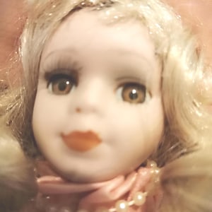 May include: A porcelain doll with blonde hair, brown eyes, and painted orange lips. The doll wears a pink fabric collar adorned with a string of small white faux pearls around the neck and a pink hat partially visible at the top of the head.