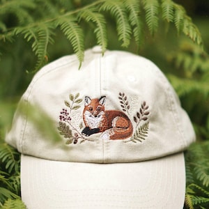 Könnte beinhalten: Eine beige Baseballkappe mit einer detaillierten Stickerei eines Rotfuchses, der zwischen grünen Blättern, braunen Zweigen und kleinen Beeren ruht. Die Kappe verfügt über eine strukturierte Krone und einen gebogenen Schirm.
