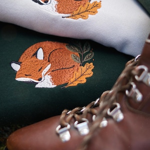 May include: Embroidered sweatshirts with a sleeping fox design. The fox is orange and white, with green leaves and an orange leaf. The sweatshirts are light green and dark green. A brown leather boot is in the foreground.