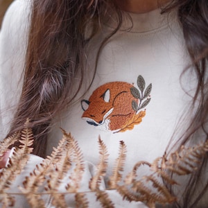 May include: A cream-coloured sweatshirt with an embroidered design of a sleeping fox in orange, brown, and white, with green leaves. The fox design is centered on the chest of the neutral-toned sweatshirt.