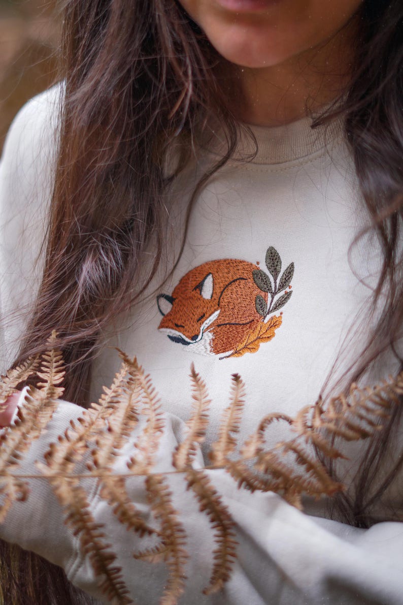 May include: A cream-coloured sweatshirt with an embroidered design of a sleeping fox in orange, brown, and white, with green leaves. The fox design is centered on the chest of the neutral-toned sweatshirt.