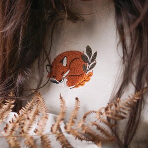 May include: Cream-coloured sweatshirt with an embroidered sleeping fox design in shades of orange, brown, and white. The fox is curled up on a bed of leaves, with a few green leaves nearby. A cosy, nature-themed apparel item.
