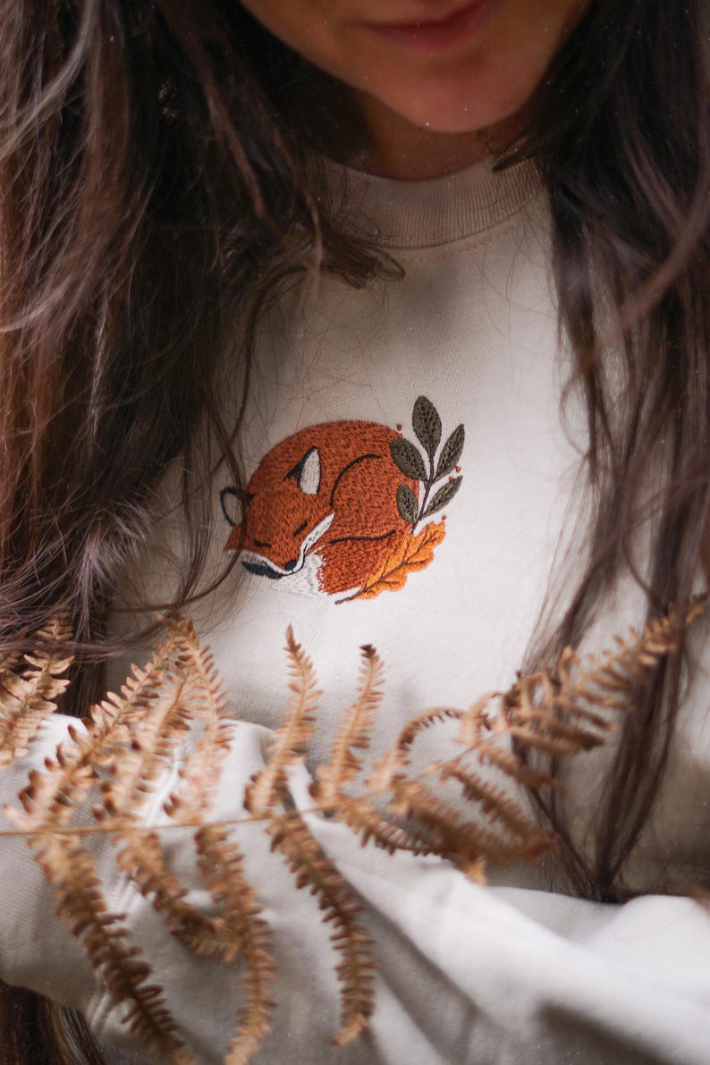 May include: Cream-coloured sweatshirt with an embroidered sleeping fox design in shades of orange, brown, and white. The fox is curled up on a bed of leaves, with a few green leaves nearby. A cosy, nature-themed apparel item.