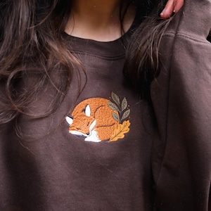 May include: A brown sweatshirt featuring an embroidered sleeping fox design. The fox is orange and white, surrounded by autumn leaves. The sweatshirt has a crew neck and is a solid colour.