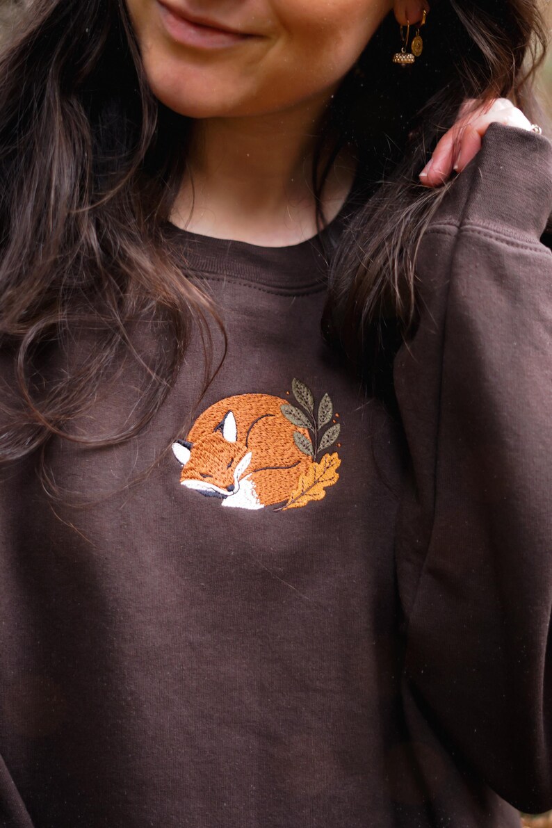 May include: A brown sweatshirt featuring an embroidered sleeping fox design. The fox is orange and white, surrounded by autumn leaves. The sweatshirt has a crew neck and is a solid colour.