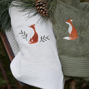 Könnte beinhalten: Zwei bestickte Handtücher, eines weiß und eines olivgrün, zeigen ein Fuchsmotiv. Der Fuchs ist in Orange- und Weißtönen dargestellt, umgeben von gestickten grünen Blättern. Ein Tannenzapfen und Tannennadeln befinden sich im Hintergrund.