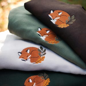 May include: Four folded sweatshirts in shades of brown, green, and white, each embroidered with a sleeping fox design. The fox is orange and white, nestled on a bed of leaves. The sweatshirts are stacked, showcasing the different colours and the detailed embroidery.