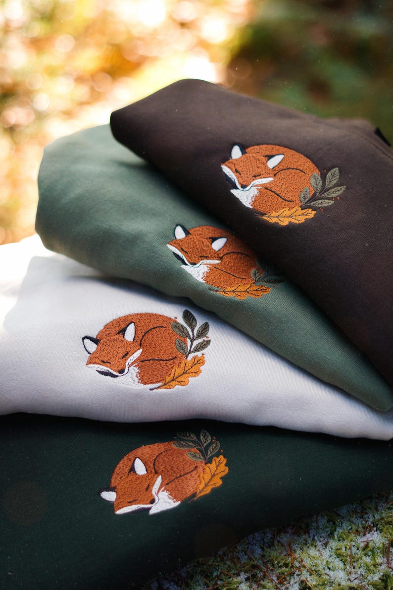 May include: Four folded sweatshirts in shades of brown, green, and white, each embroidered with a sleeping fox design. The fox is orange and white, nestled on a bed of leaves. The sweatshirts are stacked, showcasing the different colours and the detailed embroidery.