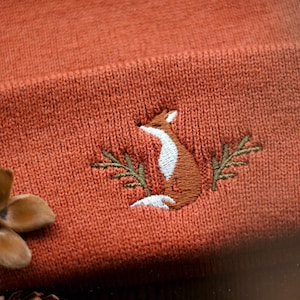 Op de afbeelding: Een close-up van een roestkleurige gebreide beanie met een geborduurd vos-ontwerp. De vos kijkt naar rechts en is omrand met wit met een bruin lichaam en groene bladeren.