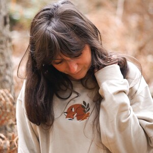 May include: A cream-coloured sweatshirt with a sleeping fox embroidery in brown and orange, accompanied by a leaf sprig. The long-sleeved top has a crew neck. The background is a blurred outdoor scene.