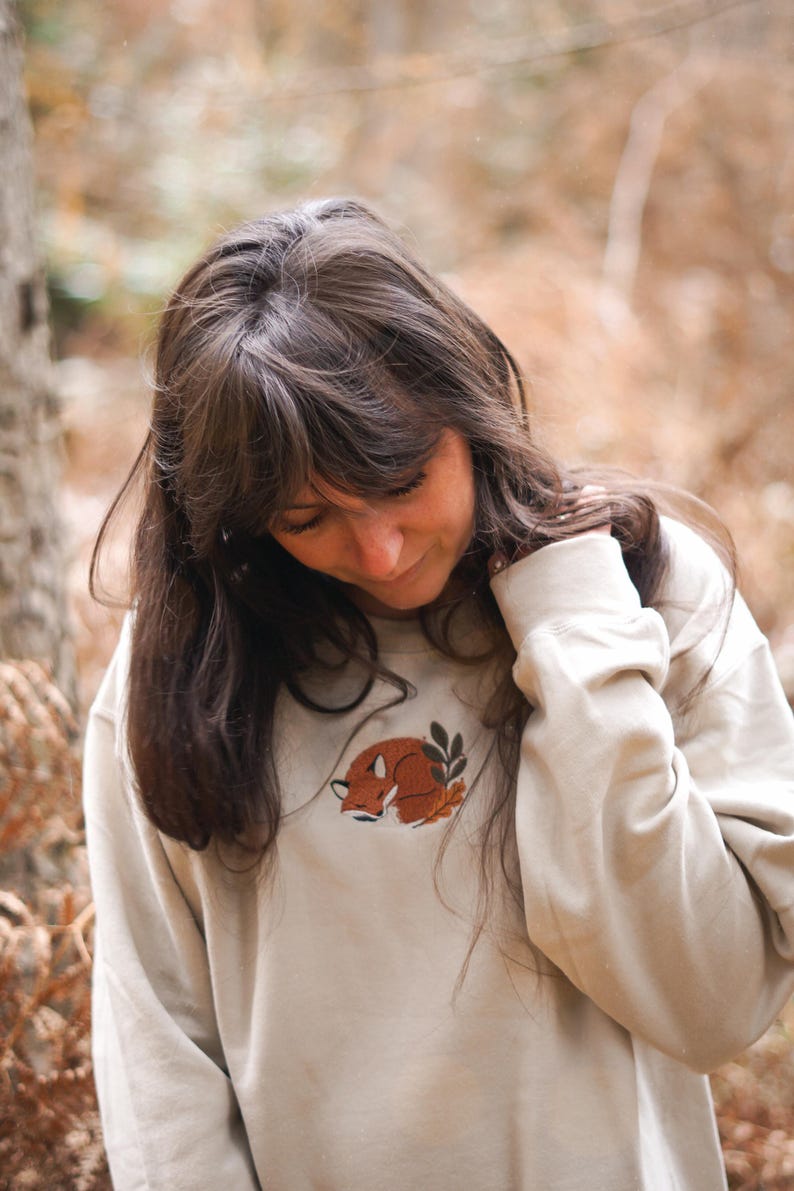 May include: A cream-coloured sweatshirt with a sleeping fox embroidery in brown and orange, accompanied by a leaf sprig. The long-sleeved top has a crew neck. The background is a blurred outdoor scene.