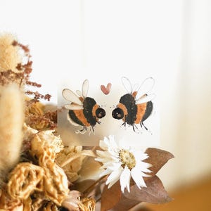 Può includere: Illustrazione ad acquerello di due api che si fronteggiano con un piccolo cuore rosa sopra di loro. Le api sono nere e arancioni con ali bianche. L'immagine è ambientata su uno sfondo di fiori secchi e uno sfondo bianco.