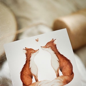 Könnte beinhalten: Zwei braune Füchse mit weißem Fell an der Brust schauen sich an. Ein kleines braunes Herz befindet sich über ihnen.