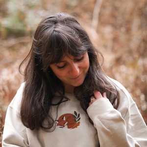 May include: A cream-coloured sweatshirt with a sleeping fox design in brown and orange, and green leaves. The sweatshirt has long sleeves and a crew neck. The person in the image is wearing the sweatshirt.