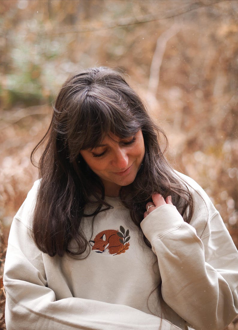 May include: A cream-coloured sweatshirt with a sleeping fox design in brown and orange, and green leaves. The sweatshirt has long sleeves and a crew neck. The person in the image is wearing the sweatshirt.