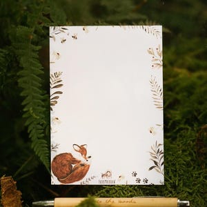 May include: A white notepad featuring a watercolour illustration of a sleeping fox, framed by floral and leaf accents. A wooden pen with the words "to the woods" is in the foreground. The notepad is set against a backdrop of green foliage.