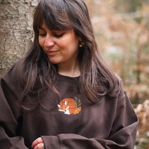 May include: A brown sweatshirt with an embroidered design of a sleeping fox. The fox is orange and white, encircled by green leaves and resting on yellow leaves. The sweatshirt is a plain brown colour.