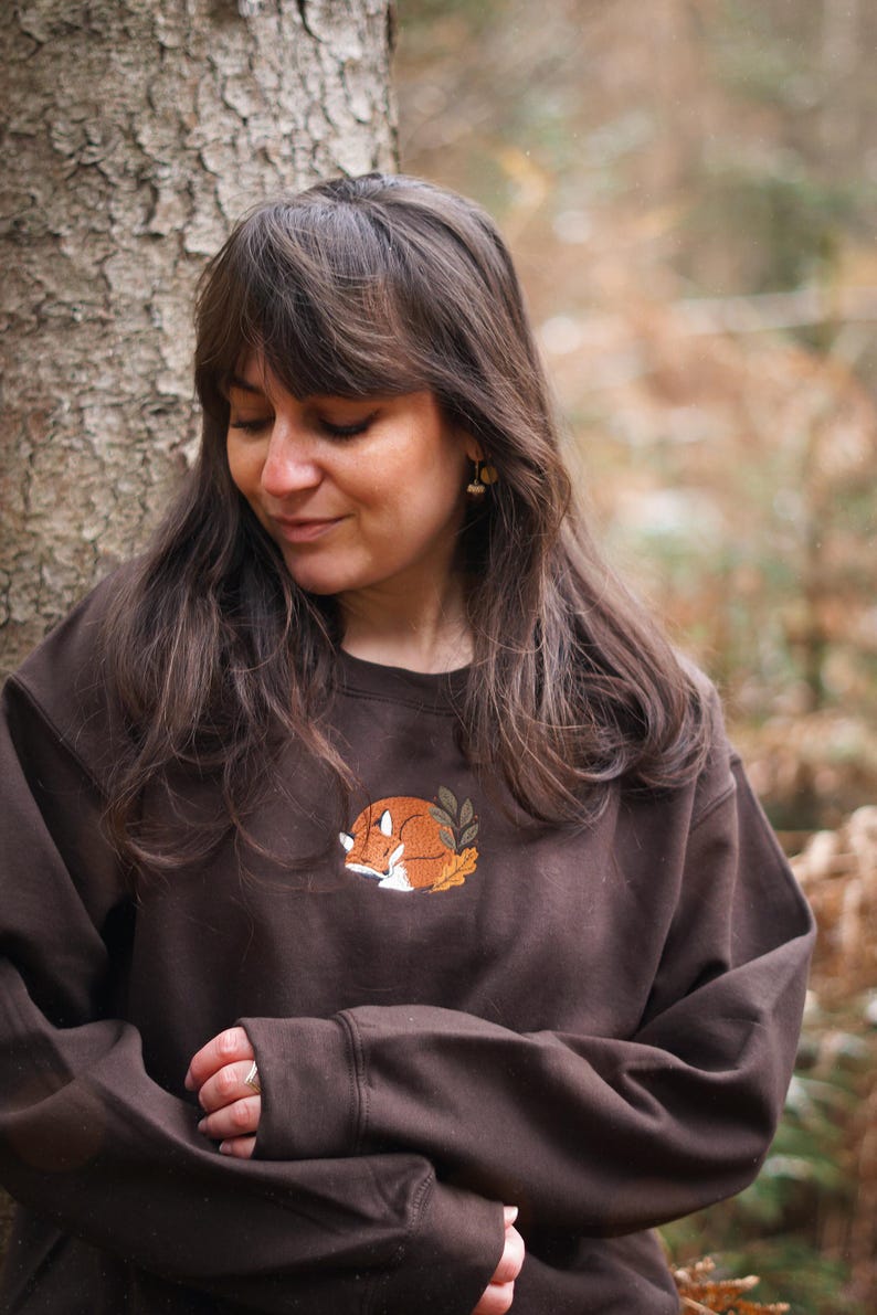May include: A brown sweatshirt with an embroidered design of a sleeping fox. The fox is orange and white, encircled by green leaves and resting on yellow leaves. The sweatshirt is a plain brown colour.