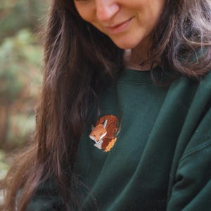 May include: A dark green sweatshirt with an embroidered fox motif. The fox is rendered in brown, orange, and white. The sweatshirt is a solid colour, offering a comfortable and casual style. The embroidery adds a touch of nature-inspired charm.