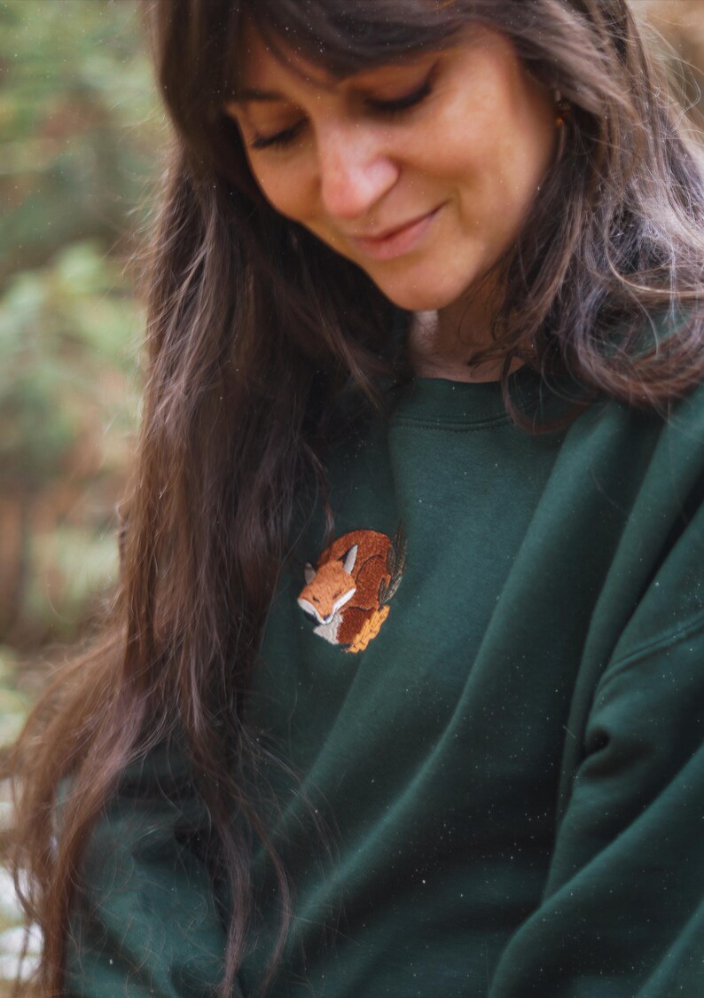 May include: A dark green sweatshirt with an embroidered fox motif. The fox is rendered in brown, orange, and white. The sweatshirt is a solid colour, offering a comfortable and casual style. The embroidery adds a touch of nature-inspired charm.