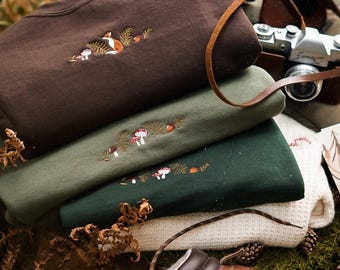 Puede incluir: Una pila de sudaderas bordadas en marrón, verde oliva y blanco, con diseños de bosque de zorros, setas y follaje. También se ven una cámara vintage y botas de senderismo de cuero, lo que sugiere un tema al aire libre.