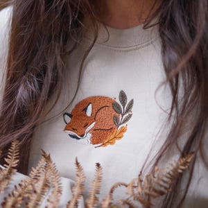 May include: Cream-coloured sweatshirt with an embroidered sleeping fox design. The fox is in shades of brown and orange, surrounded by leaves. The sweatshirt is a neutral colour, suitable for casual wear.