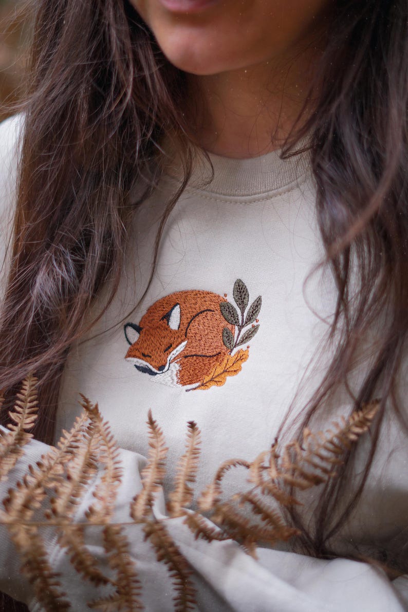 May include: Cream-coloured sweatshirt with an embroidered sleeping fox design. The fox is in shades of brown and orange, surrounded by leaves. The sweatshirt is a neutral colour, suitable for casual wear.