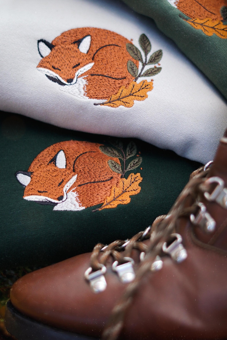 May include: Embroidered sweatshirts with a sleeping fox design. The fox is orange and white, with green leaves and an orange leaf. The sweatshirts are light green and dark green. A brown leather boot is in the foreground.