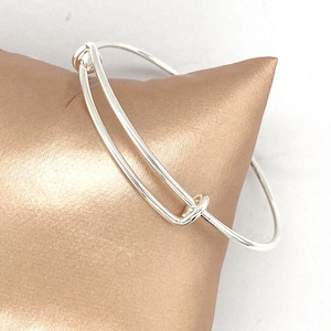 May include: A silver bangle bracelet with a double loop design. The bracelet is open and has a simple, elegant style.