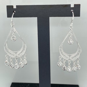 May include: Silver dangle earrings with a teardrop design and clear crystal accents. The earrings have a filigree pattern and a dangling chain with multiple crystals.