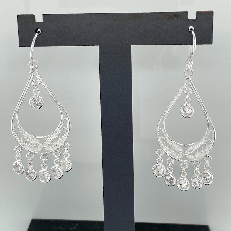 May include: Silver dangle earrings with a teardrop design and clear crystal accents. The earrings have a filigree pattern and a dangling chain with multiple crystals.