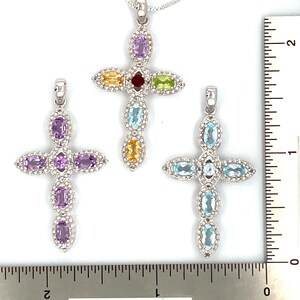 Puede incluir: Tres colgantes de cruz de plata con piedras preciosas de diferentes colores. La cruz superior tiene una piedra roja, amarilla, verde y morada. La cruz del medio tiene piedras moradas. La cruz inferior tiene piedras azules.