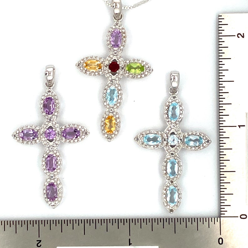 Puede incluir: Tres colgantes de cruz de plata con piedras preciosas de diferentes colores. La cruz superior tiene una piedra roja, amarilla, verde y morada. La cruz del medio tiene piedras moradas. La cruz inferior tiene piedras azules.