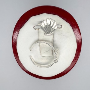 May include: Two silver rings with a seashell design. The rings are displayed on a red velvet ring holder.