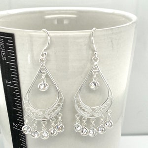 May include: Silver dangle earrings with a teardrop design and clear crystal accents. The earrings have a filigree pattern and a dangling row of smaller crystals.