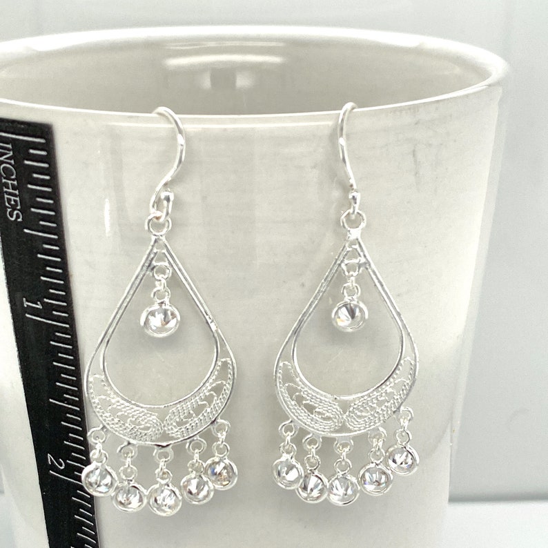 May include: Silver dangle earrings with a teardrop design and clear crystal accents. The earrings have a filigree pattern and a dangling row of smaller crystals.