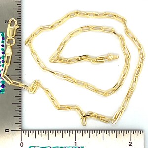 K&ouml;nnte beinhalten: Eine goldene Kettenkette mit einem Boxverschluss. Die Kette besteht aus kleinen, miteinander verbundenen ovalen Formen. Die Kette liegt auf einer wei&szlig;en Oberfl&auml;che mit einem Lineal als Ma&szlig;stab.