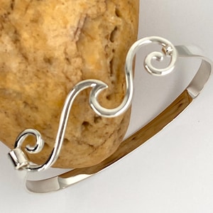 May include: Silver wave bangle bracelet with a delicate, swirling design. The bracelet is open and adjustable to fit most wrists.