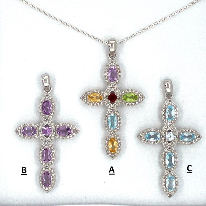 Puede incluir: Tres colgantes de cruz de plata con piedras preciosas de diferentes colores. El colgante superior tiene una piedra preciosa roja, verde, amarilla y azul. El colgante izquierdo tiene piedras preciosas moradas. El colgante derecho tiene piedras preciosas azules. Cada colgante tiene una cadena de plata.