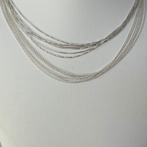 Puede incluir: Un collar plateado de capas con m&uacute;ltiples cadenas de diferentes anchos. El collar se muestra en un busto de maniqu&iacute; blanco.