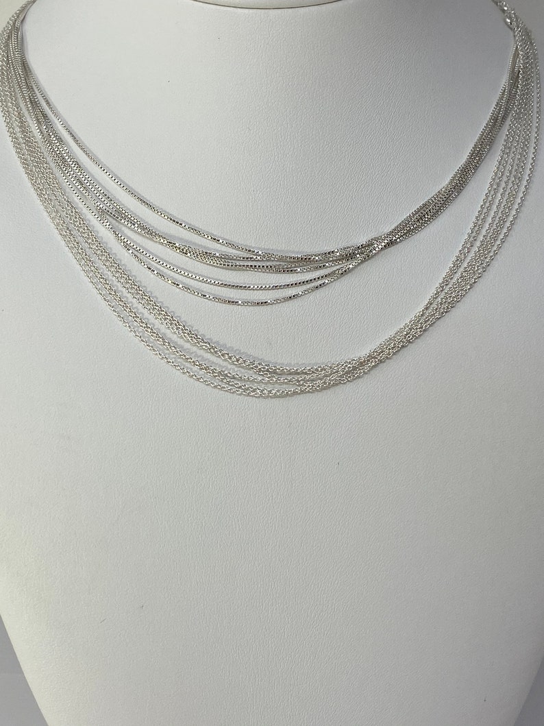 Puede incluir: Un collar plateado de capas con m&uacute;ltiples cadenas de diferentes anchos. El collar se muestra en un busto de maniqu&iacute; blanco.
