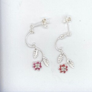 May include: A pair of silver-coloured drop earrings. Each earring has a curved vine design with leaves and a red flower with a clear centre. The earrings have a post-back fastening.