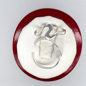 May include: A silver ring with a wavy design. The ring is displayed on a red and white display stand.