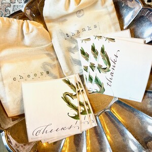 May include: A silver tray with two white fabric bags with the words "thanks" and "cheers" printed on them. There are also several white cards with green floral designs and the words "thanks" and "cheers" written in calligraphy.