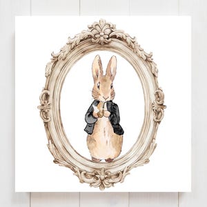 Puede incluir: Ilustración en acuarela de un conejo con una chaqueta negra sosteniendo un objeto pequeño, enmarcada dentro de un marco ovalado adornado de color beige. La obra de arte está sobre un fondo blanco, evocando una estética de cuento de hadas clásica.