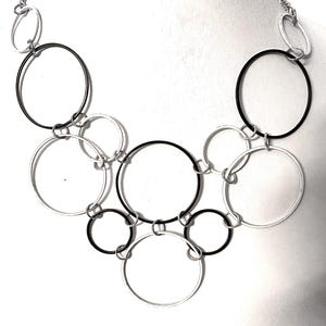 May include: A silver-toned necklace featuring a series of interconnected circles in varying sizes. Some circles are black, others are silver, creating a modern, geometric design. The necklace is displayed against a white background.